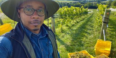 photo of a man with a hat standing in front of a vineyard